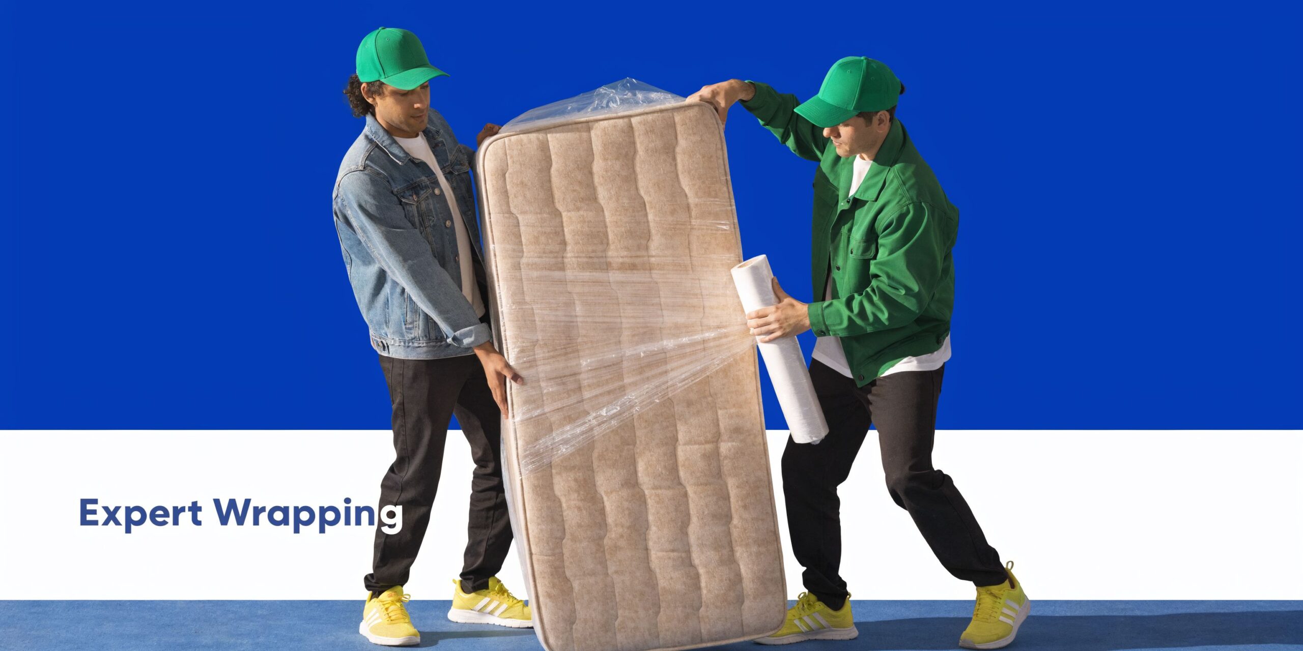 Two movers in green caps wrapping a mattress in clear plastic film against a blue background.