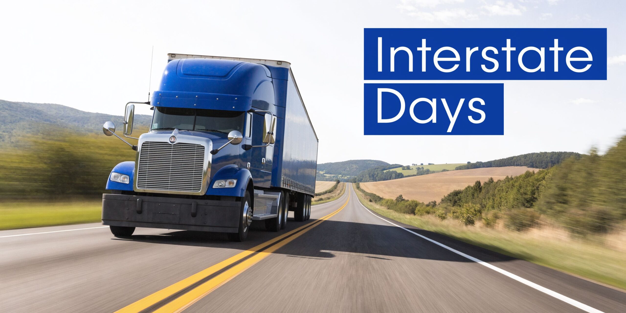 A blue semi truck driving down an empty highway against a scenic countryside background at sunset.